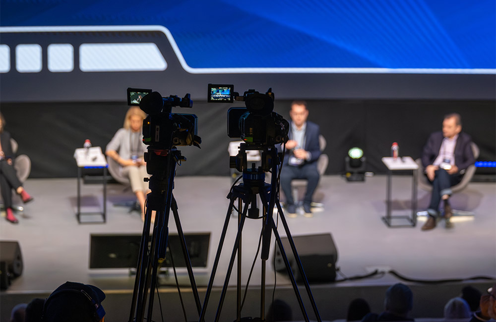 Two professional video cameras on tripods capture a live panel discussion during a hybrid event.